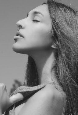 A black and white portrait of a person in profile with closed eyes, exuding a sense of calm and introspection. The individual has long hair and is holding onto a piece of clothing at the shoulder.
