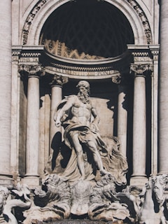 A classical stone sculpture depicting a powerful male figure, possibly a deity, surrounded by an ornate architectural setting. The scene includes flowing robes and dynamic, carved elements giving a sense of movement and grandeur. Horses and intricate designs complement the central statue, all crafted in a detailed and realistic manner.