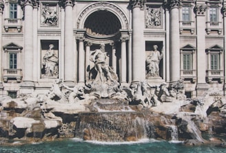 A grand fountain adorned with intricate sculptures and statues, surrounded by an ornate architectural facade featuring columns and decorative reliefs. Water cascades over sculpted rocks into a turquoise pool below, adding a sense of movement and life to the scene.