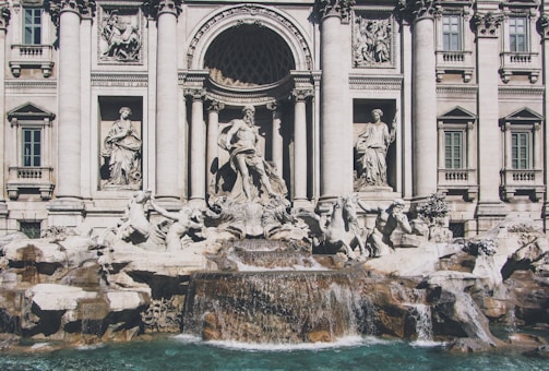 A grand fountain adorned with intricate sculptures and statues, surrounded by an ornate architectural facade featuring columns and decorative reliefs. Water cascades over sculpted rocks into a turquoise pool below, adding a sense of movement and life to the scene.