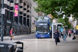 A busy urban street scene with a blue double-decker bus marked '163' heading to Castleford. People are walking on the sidewalk, some looking at their phones or in conversation. A man in a red shirt is crossing the street. The area is lined with tall buildings, and there are signs and advertisements on lamp posts and bus stops. Trees have green foliage, adding some natural contrast to the urban environment.