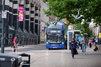 A busy urban street scene with a blue double-decker bus marked '163' heading to Castleford. People are walking on the sidewalk, some looking at their phones or in conversation. A man in a red shirt is crossing the street. The area is lined with tall buildings, and there are signs and advertisements on lamp posts and bus stops. Trees have green foliage, adding some natural contrast to the urban environment.