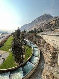 A winding concrete water channel flows through a green landscape, bordered by tall, lush trees. In the background, a rugged mountain rises steeply, with a clear, blue sky overhead. There is a road running parallel to the channel and a parking area occupied by several cars.