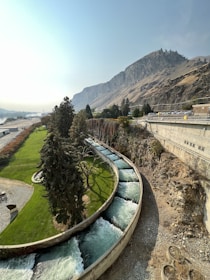 A winding concrete water channel flows through a green landscape, bordered by tall, lush trees. In the background, a rugged mountain rises steeply, with a clear, blue sky overhead. There is a road running parallel to the channel and a parking area occupied by several cars.