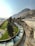 A winding concrete water channel flows through a green landscape, bordered by tall, lush trees. In the background, a rugged mountain rises steeply, with a clear, blue sky overhead. There is a road running parallel to the channel and a parking area occupied by several cars.