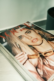 A magazine cover features a close-up image of a person with long hair, holding sunglasses. The magazine appears to be on a desk or table, with part of the title 'Vogue' visible.
