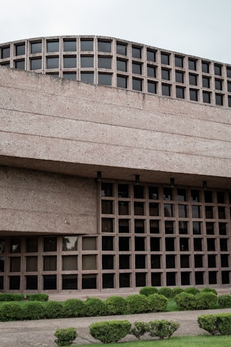 A modern architectural building features a grid-like pattern of square windows. The facade is made of textured concrete, giving it a robust and industrial appearance. The lower portion of the building is complemented by neatly trimmed hedges and green grass, enhancing the overall aesthetic.