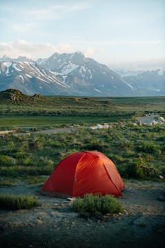 A vibrant red tent is set up in a natural landscape, surrounded by green grass and small bushes. The backdrop showcases majestic, snow-capped mountains under a clear, blue sky.