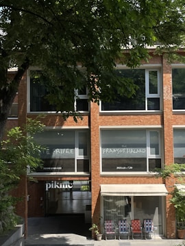 A brick building with large windows displaying text. The upper windows feature handwritten-style text promoting an exhibition by François Halard with dates. An entrance labeled 'piknic' is below and leads into an open space inside. Three folding chairs are arranged side by side under an awning next to potted plants. Lush green trees frame the upper part of the image, casting shadows on the building.