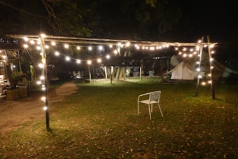 A nighttime outdoor scene with a string of warm white lights hanging on a wooden framework. There is a single white chair on a grassy area. In the background, tents are partially visible among trees, suggesting a campsite or garden setting.