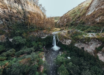 A picturesque waterfall cascades into a dark pool surrounded by lush greenery and steep rocky cliffs. The landscape is rich with dense vegetation and natural beauty, creating a serene and untouched atmosphere.