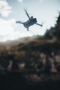 A drone with four propellers is flying in mid-air against a backdrop of a blurred landscape. The sky is partly cloudy, with soft, diffused lighting creating a moody atmosphere.