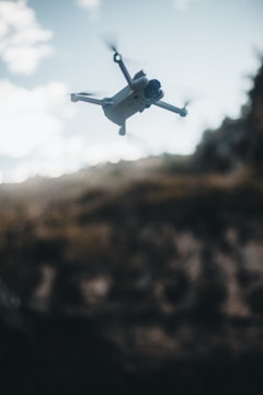 A drone with four propellers is flying in mid-air against a backdrop of a blurred landscape. The sky is partly cloudy, with soft, diffused lighting creating a moody atmosphere.