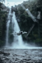 A drone hovers in front of a tall, cascading waterfall set within a lush, green forest. The water appears to be in motion, creating a misty effect at the base, while the forest background is slightly out of focus, giving depth to the image.