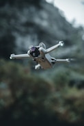 A compact drone with four propellers hovers in mid-air, framed against a blurred natural background with greenery. The drone's design appears sleek and modern, equipped with a camera and lights on its arms.