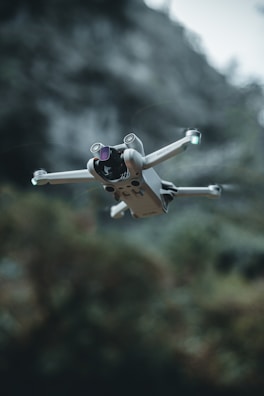 A compact drone with four propellers hovers in mid-air, framed against a blurred natural background with greenery. The drone's design appears sleek and modern, equipped with a camera and lights on its arms.