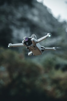 A compact drone with four propellers hovers in mid-air, framed against a blurred natural background with greenery. The drone's design appears sleek and modern, equipped with a camera and lights on its arms.