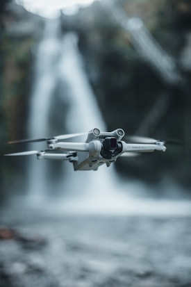 A drone hovers mid-air with a blurred waterfall in the background. The drone is light gray and detailed with visible propellers. The natural surroundings include flowing water and soft focus foliage.