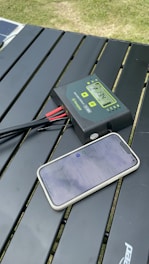 A digital device with cables displays a reading on a screen, placed on a slatted outdoor table beside a smartphone showing a web page. The setting is outdoors with visible grass in the background.