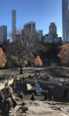 A bustling urban park scene with people enjoying recreational activities. The foreground features a rocky area and playground with a slide. People are walking and gathering around, and the trees have autumn foliage. Skyscrapers line the background under a clear blue sky.