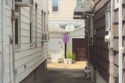 A narrow alley between two residential buildings with siding. An air conditioning unit is mounted on each side. At the end of the alley, a purple shirt is hanging on a line. There are potted plants and a brown door visible in the background.