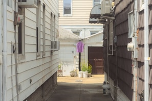 A narrow alley between two residential buildings with siding. An air conditioning unit is mounted on each side. At the end of the alley, a purple shirt is hanging on a line. There are potted plants and a brown door visible in the background.