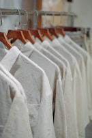 A row of white shirts neatly arranged on wooden hangers in a closet or store setting. The shirts have a simple design, creating a clean and organized appearance.