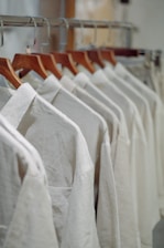 A row of white shirts neatly arranged on wooden hangers in a closet or store setting. The shirts have a simple design, creating a clean and organized appearance.