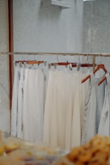 Several pieces of light-colored clothing are hanging on a metal clothing rack. The garments are displayed against a minimally textured wall with a reflective surface on the right. The scene has a soft, neutral tone with a focus on simplicity and calmness.