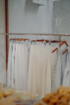 Several pieces of light-colored clothing are hanging on a metal clothing rack. The garments are displayed against a minimally textured wall with a reflective surface on the right. The scene has a soft, neutral tone with a focus on simplicity and calmness.