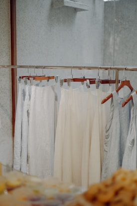Several pieces of light-colored clothing are hanging on a metal clothing rack. The garments are displayed against a minimally textured wall with a reflective surface on the right. The scene has a soft, neutral tone with a focus on simplicity and calmness.