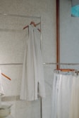 A minimalist setting featuring an elegant, sleeveless white dress hanging on a wooden hanger against a speckled wall. Below it, several garments in similar light hues are displayed on hangers, creating a serene and harmonious arrangement.
