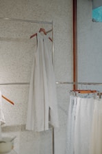 A minimalist setting featuring an elegant, sleeveless white dress hanging on a wooden hanger against a speckled wall. Below it, several garments in similar light hues are displayed on hangers, creating a serene and harmonious arrangement.