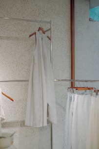 A minimalist setting featuring an elegant, sleeveless white dress hanging on a wooden hanger against a speckled wall. Below it, several garments in similar light hues are displayed on hangers, creating a serene and harmonious arrangement.