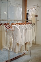 A row of beige and light-colored clothing items on wooden hangers displayed on a metal rack in a store setting. The background contains other racks with dresses that appear sheer and slightly more formal. The setting is minimalistic with a calm and neutral aesthetic.