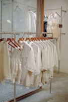 A row of beige and light-colored clothing items on wooden hangers displayed on a metal rack in a store setting. The background contains other racks with dresses that appear sheer and slightly more formal. The setting is minimalistic with a calm and neutral aesthetic.
