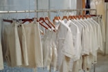 A row of clothing items hangs neatly on wooden hangers along a metal clothing rack. The garments are composed of light-colored, possibly linen fabric, including shirts, shorts, and overalls. The setting appears to be inside a clothing store or boutique with a minimalistic and organized display.