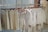 A row of clothing items hangs neatly on wooden hangers along a metal clothing rack. The garments are composed of light-colored, possibly linen fabric, including shirts, shorts, and overalls. The setting appears to be inside a clothing store or boutique with a minimalistic and organized display.