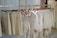 A row of clothing items hangs neatly on wooden hangers along a metal clothing rack. The garments are composed of light-colored, possibly linen fabric, including shirts, shorts, and overalls. The setting appears to be inside a clothing store or boutique with a minimalistic and organized display.