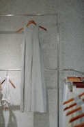 A simple, sleeveless dress made of a light fabric hangs on a wooden hanger against a speckled wall. Other clothing items and hangers can be seen to the side, suggesting a minimalistic and organized setting.