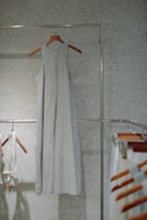 A simple, sleeveless dress made of a light fabric hangs on a wooden hanger against a speckled wall. Other clothing items and hangers can be seen to the side, suggesting a minimalistic and organized setting.