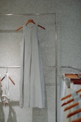 A simple, sleeveless dress made of a light fabric hangs on a wooden hanger against a speckled wall. Other clothing items and hangers can be seen to the side, suggesting a minimalistic and organized setting.