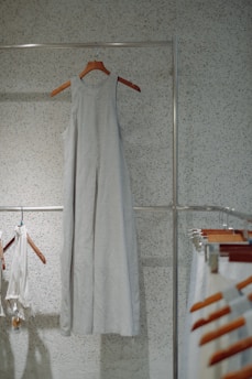 A simple, sleeveless dress made of a light fabric hangs on a wooden hanger against a speckled wall. Other clothing items and hangers can be seen to the side, suggesting a minimalistic and organized setting.