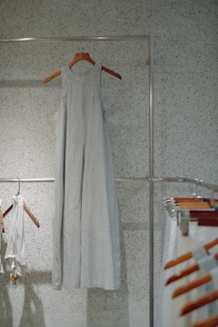 A simple, sleeveless dress made of a light fabric hangs on a wooden hanger against a speckled wall. Other clothing items and hangers can be seen to the side, suggesting a minimalistic and organized setting.