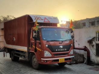 A brown delivery truck with decorative elements is parked on a paved area near a wall. The truck has various inscriptions and a yellow license plate, and the sun is setting in the background, giving a warm glow.