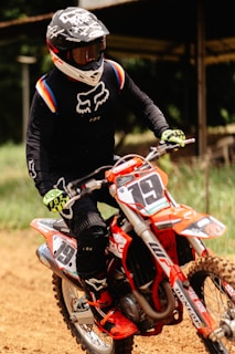 A person is riding a motocross bike on a dirt track. They are wearing a black racing outfit with colorful accents, protective gloves, and boots. The rider's helmet is black with white designs and a visor. The bike is primarily orange with visible numbers and sponsor decals.