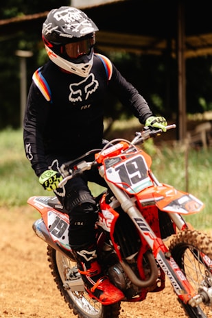 A person is riding a motocross bike on a dirt track. They are wearing a black racing outfit with colorful accents, protective gloves, and boots. The rider's helmet is black with white designs and a visor. The bike is primarily orange with visible numbers and sponsor decals.