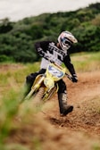 A motorcyclist navigating a dirt track on a motocross bike. The person is wearing full protective gear, including a helmet and a black and white jersey. The environment is natural, with grass and greenery in the background along a winding dirt path.