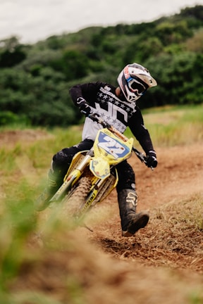 A motorcyclist navigating a dirt track on a motocross bike. The person is wearing full protective gear, including a helmet and a black and white jersey. The environment is natural, with grass and greenery in the background along a winding dirt path.