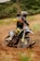 A motocross rider aggressively leans into a turn on a dirt track. The rider is wearing protective gear including a helmet and colorful suit, and the motorcycle's wheels are kicking up dirt as it speeds through the curve. The background features lush greenery, suggesting an outdoor setting.